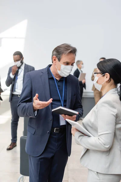 Interracial business people in medical masks, with notebook and digital tablet, talking in hall during conference — Stock Photo