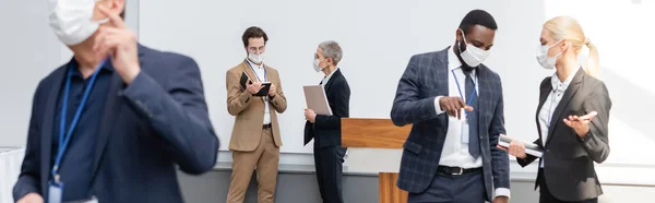 Uomo d'affari afroamericano in maschera medica che punta con dito vicino collega e partner, striscione — Foto stock