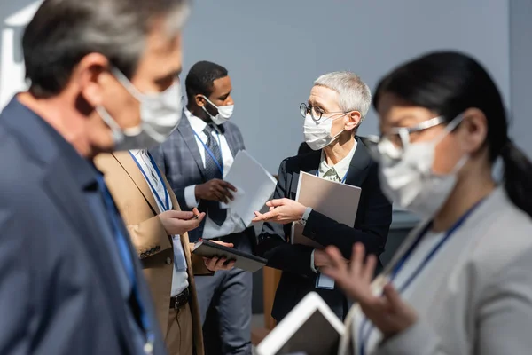 Mature businesswoman and african american businessman in medical masks talking near blurred interracial colleagues — Stock Photo
