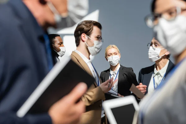 Багатонаціональні бізнесмени в медичних масках говорять під час перерви на семінарі, розмитий передній план — стокове фото