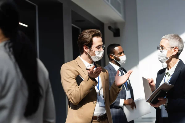 Young businessman in medical mask gesturing near mature businesswoman and interracial colleagues — Stock Photo