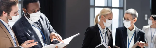 African american businessman in medical mask pointing at documents near young colleague, banner — Stock Photo