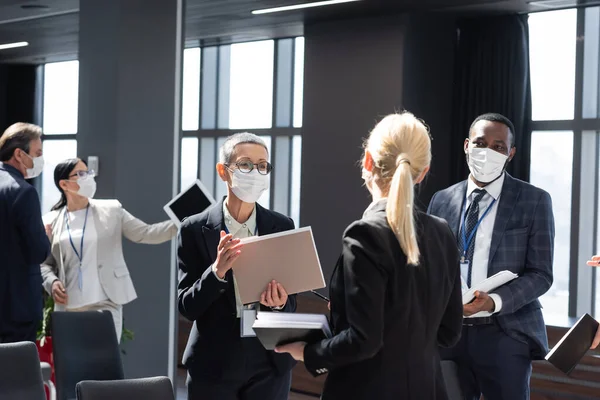 Зріла бізнес-леді в медичній масці тримає документи під час розмови з міжрасовими колегами — стокове фото