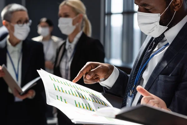 Hombre de negocios afroamericano en máscara médica apuntando a documentos con gráficos cerca de colegas borrosos - foto de stock