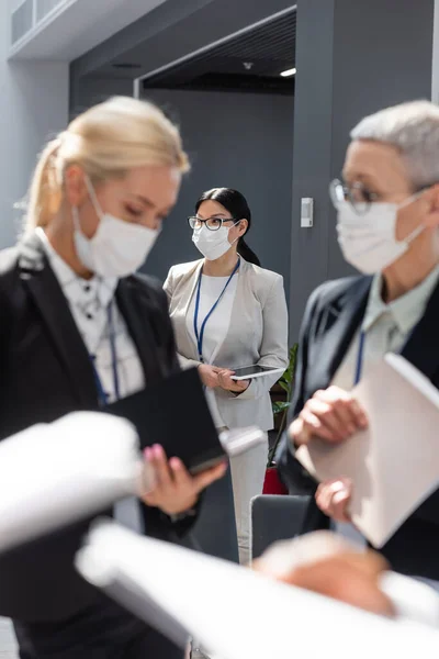Вибірковий фокус азіатської бізнес-леді в медичній масці біля колег, які говорять на розмитому передньому плані — стокове фото