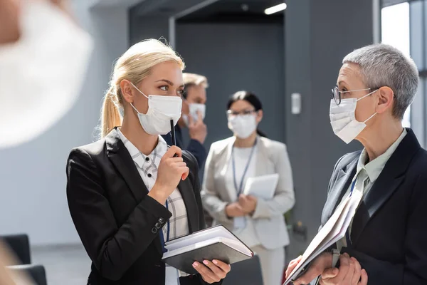 Молода бізнес-леді в медичній масці тримає ручку і блокнот біля зрілого колеги — стокове фото