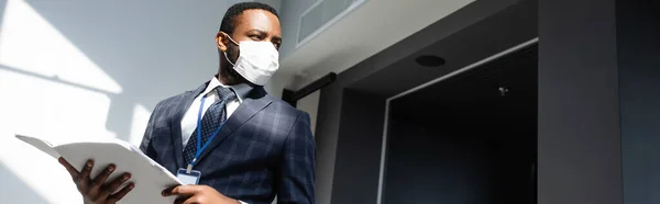 Homme d'affaires afro-américain en masque médical tenant des documents tout en regardant loin dans le bureau, bannière — Photo de stock