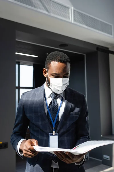 Africano americano hombre de negocios en médico máscara celebración carpeta con documentos en la oficina - foto de stock