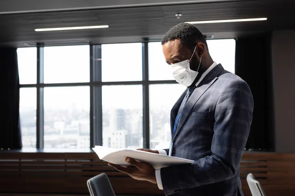 Hombre de negocios afroamericano en máscara protectora mirando documentos en la oficina - foto de stock
