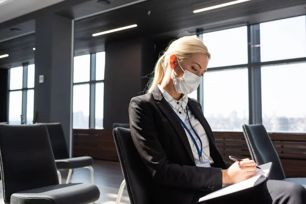 Femme d'affaires en masque médical écrit dans un cahier tout en étant assis dans la salle de conférence — Photo de stock