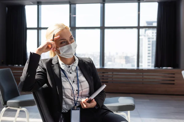 Giovane donna d'affari in maschera di sicurezza guardando lontano mentre seduto in sala conferenze con notebook — Foto stock