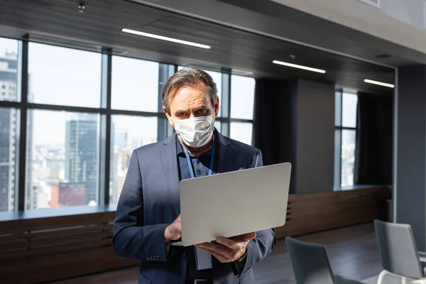 Homem de negócios de meia idade em máscara médica usando laptop enquanto estava no escritório — Fotografia de Stock