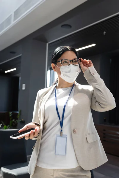 Asiática empresária segurando celular e ajustando óculos enquanto olha para longe no escritório — Fotografia de Stock