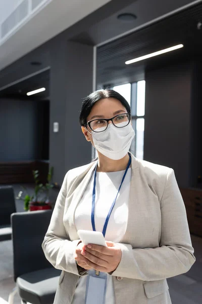 Asian businesswoman in medical mask and eyeglasses holding smartphone in office — Stock Photo