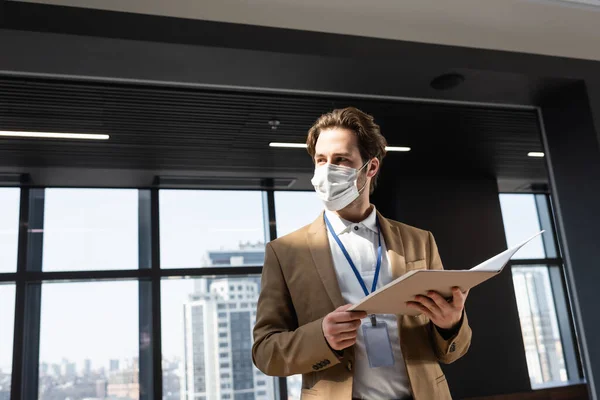 Homem de negócios em máscara de segurança segurando documentos enquanto olha para longe no escritório — Fotografia de Stock