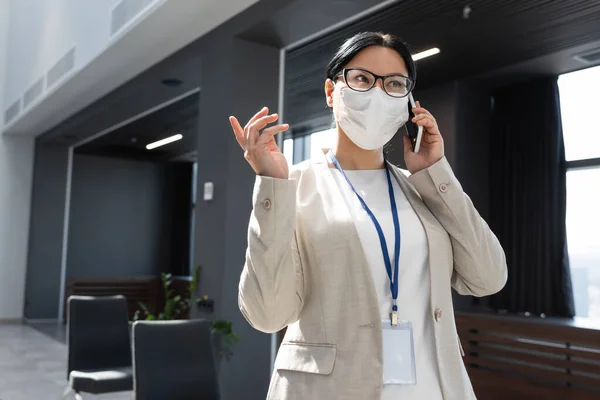 Mujer de negocios asiática en máscara médica señalando con el dedo mientras habla por teléfono móvil en la oficina - foto de stock