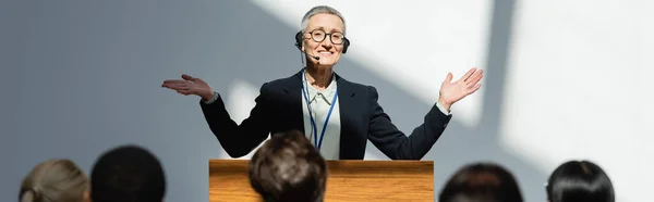 Lächelnder Dozent, der während des Seminars mit offenen Armen vor verschwommenen Teilnehmern steht, Banner — Stockfoto