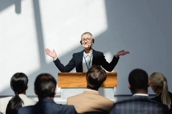 Smiling lecturer standing with open arms during conference near participants on blurred foreground — Stock Photo