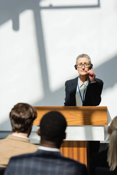 Mature speaker adjusting headset during seminar with business people on blurred foreground — Stock Photo