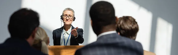 Altavoz de mediana edad que apunta a la gente de negocios borrosa durante el seminario, pancarta - foto de stock