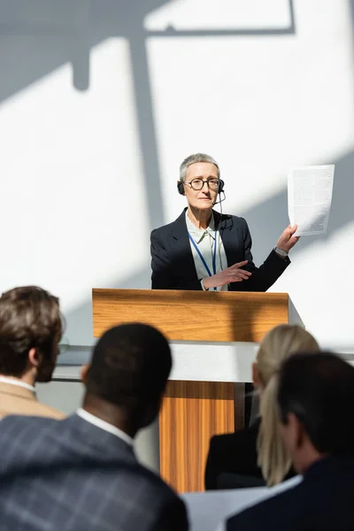 Orador de mediana edad señalando documento cerca de audiencia borrosa durante conferencia de negocios - foto de stock