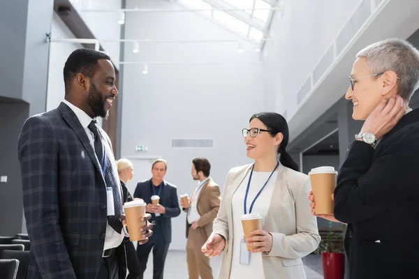 Des hommes d'affaires multiethniques souriants tenant un café à emporter tout en parlant pendant la conférence — Photo de stock