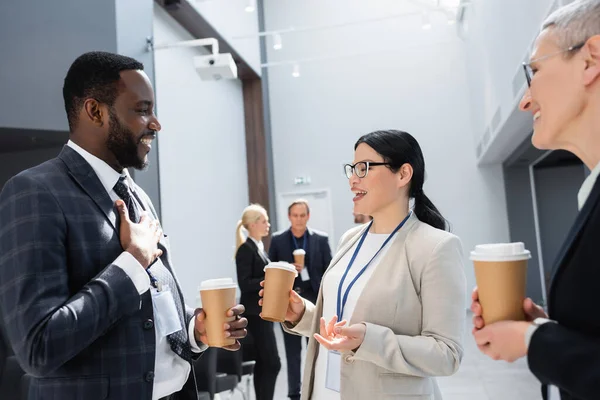 Smiling multiethnic business colleagues with coffee to go talking during meeting — Stock Photo