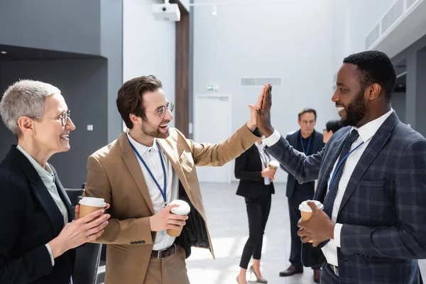 Hommes d'affaires multiethniques donnant haute cinq tout en tenant des tasses de papier pendant la pause café — Photo de stock