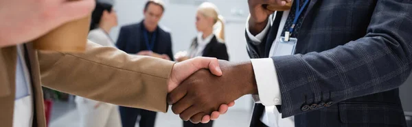 Interracial businessmen shaking hands near colleagues on blurred background, banner — Stock Photo