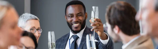 Sonriente hombre de negocios afroamericano sosteniendo copa de champán cerca de colega en primer plano borrosa, bandera - foto de stock