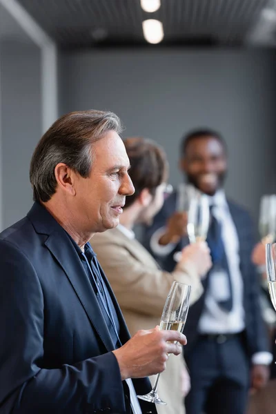 Mature businessman holding champagne glass near colleagues on blurred background — Stock Photo