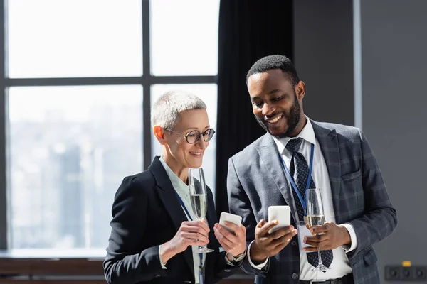 Socios comerciales interracial feliz con champán y teléfonos celulares hablando durante la conferencia de negocios - foto de stock