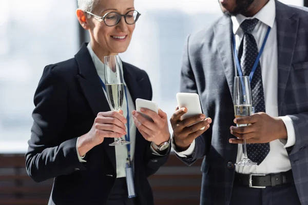 Socios comerciales interracial celebración de champán y teléfonos inteligentes durante la conversación - foto de stock