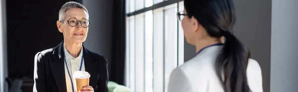 Reife Geschäftsfrau hält Kaffee in der Hand, um sich Kollegin auf verschwommenem Vordergrund zu nähern, Banner — Stockfoto