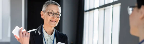 Mature businesswoman with smartphone talking to blurred colleague during conference, banner — Stock Photo