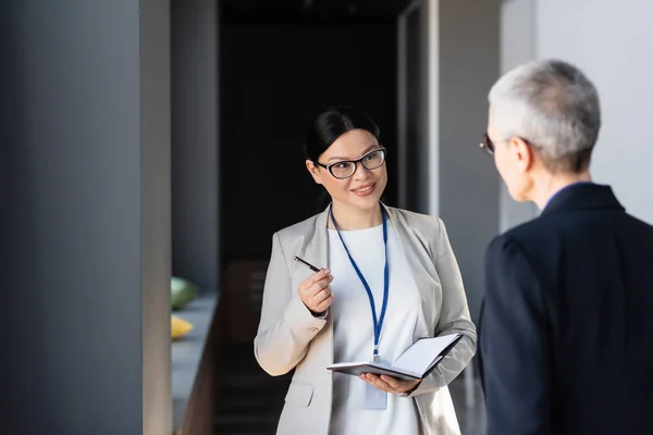 Asiático mujer de negocios con notebook y pluma hablando con colega en borrosa primer plano - foto de stock