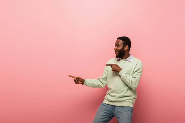 Vista lateral do homem afro-americano sorrindo e apontando para o fundo rosa — Fotografia de Stock