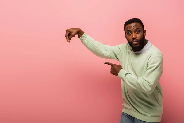 Afro-américain homme regardant caméra tout en tenant quelque chose et pointant du doigt sur fond rose — Photo de stock