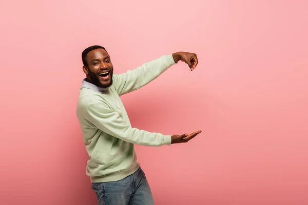 Homme afro-américain positif pointant avec la main et tenant quelque chose sur fond rose — Photo de stock