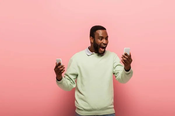 Alegre hombre afroamericano sosteniendo teléfonos inteligentes sobre fondo rosa - foto de stock