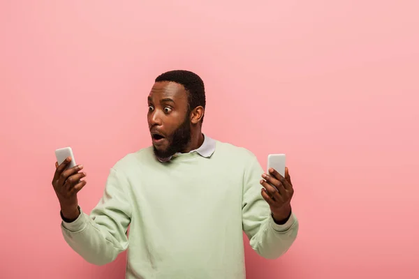 Hombre afroamericano asombrado sosteniendo teléfonos celulares sobre fondo rosa - foto de stock