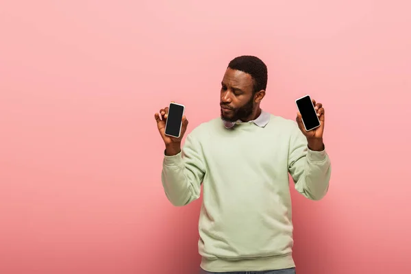 African american man holding smartphones with blank screen on pink background — Stock Photo