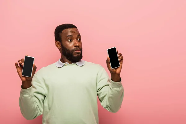 Impresionado hombre afroamericano sosteniendo teléfonos inteligentes sobre fondo rosa - foto de stock