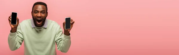 Emocionado hombre afroamericano con teléfonos inteligentes aislados en rosa, bandera - foto de stock