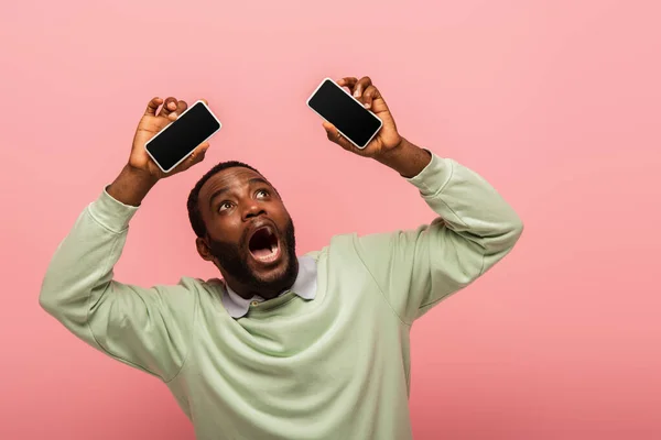 Hombre afroamericano asustado sosteniendo teléfonos celulares con pantalla en blanco aislado en rosa - foto de stock