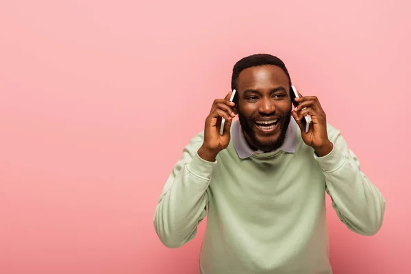 Sonriente hombre afroamericano hablando en teléfonos inteligentes aislados en rosa - foto de stock
