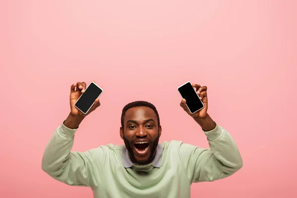 African american man holding smartphones with blank screen isolated on pink — Stock Photo