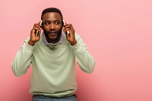 Joven afroamericano hablando en teléfonos móviles sobre fondo rosa - foto de stock