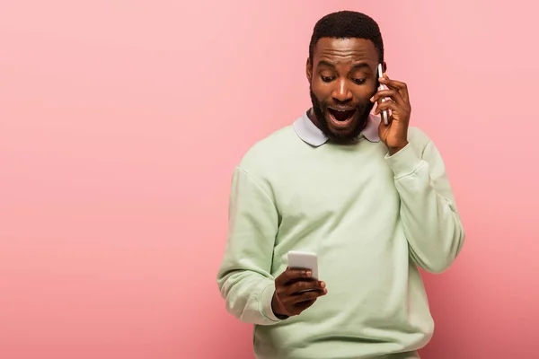 Hombre afroamericano asombrado hablando en teléfono inteligente y utilizando el teléfono celular en el fondo rosa - foto de stock