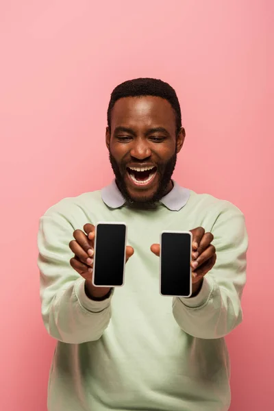 Positive afrikanisch-amerikanische Mann hält Smartphones auf verschwommenem Vordergrund isoliert auf rosa — Stockfoto